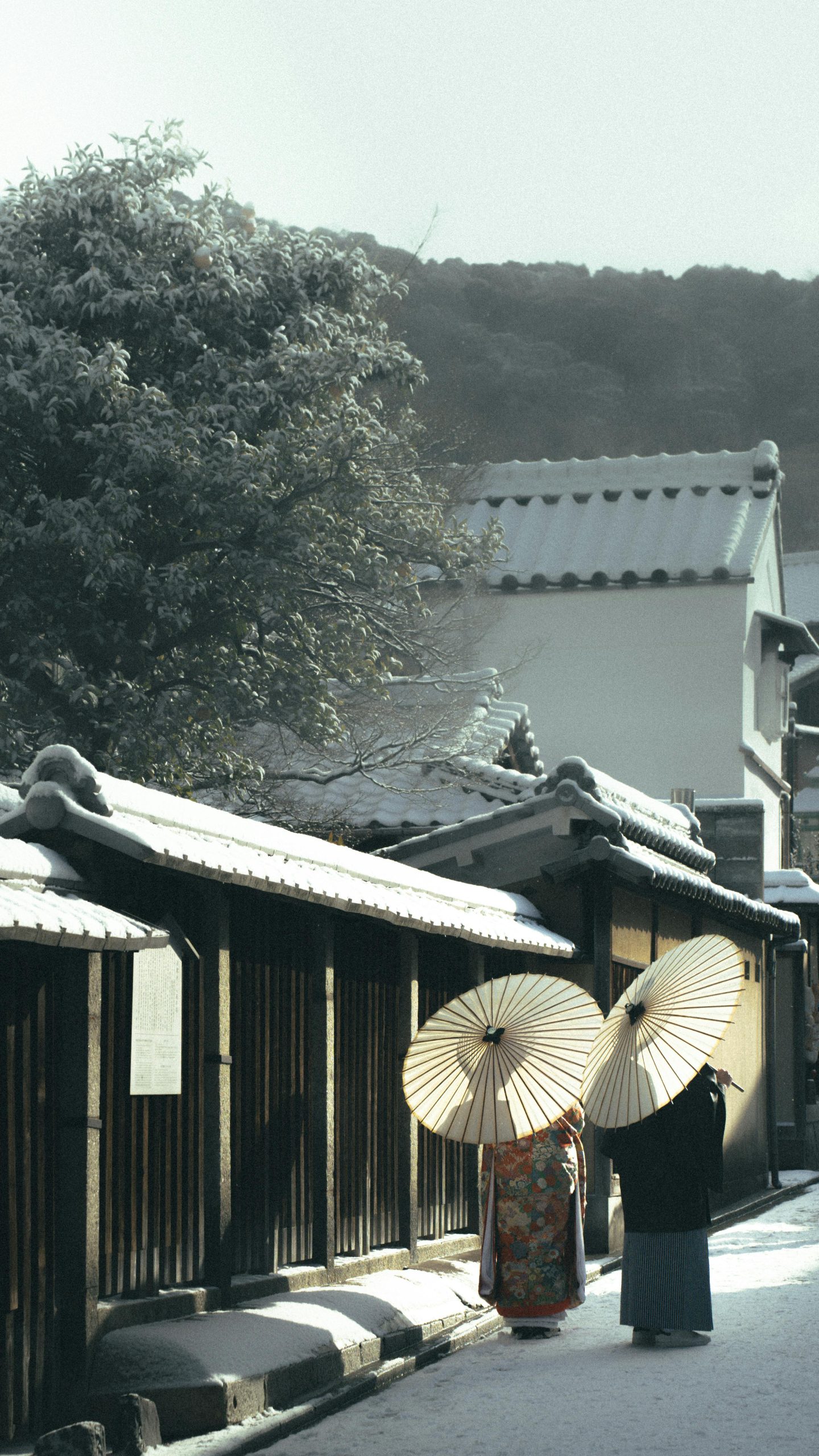 Two people in traditional attire with umbrellas stroll a snowy street in Japan.